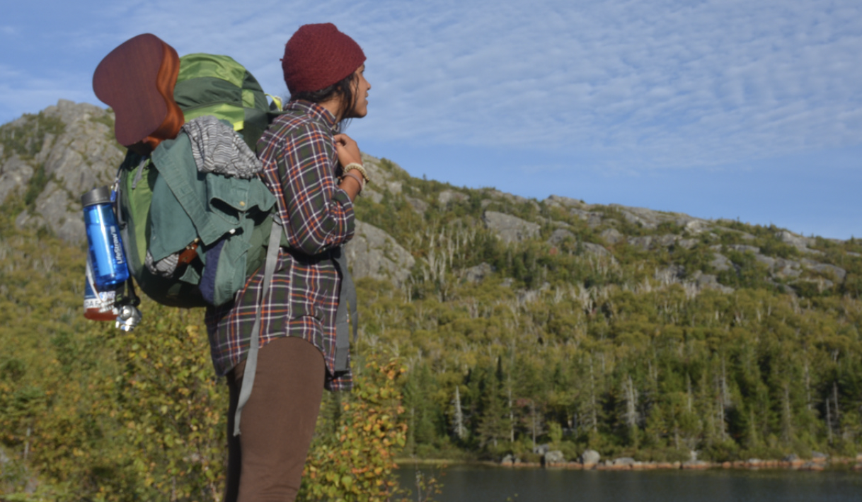 Person wearing a backpack standing in the wilderness near water and granite rocks.
